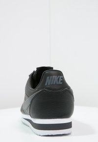 Baskets en cuir noir à finition texturée, avec une semelle intermédiaire blanche et un logo Nike gris sur le talon, design classique de chaussure de course.