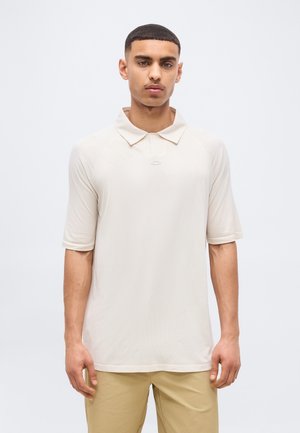 Mladý muž s krátkymi vlasmi oblečený v svetlobéžovej krátkosvejovej polokošeli a kaki nohaviciach stojaci pred jednoduchým bielym pozadím.