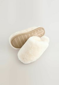 Kreminių spalvų šlepetės be užsegimo, pagamintos iš minkštos vilnos, su tekstūruota smėlio spalvos guma padais, apvalios formos.