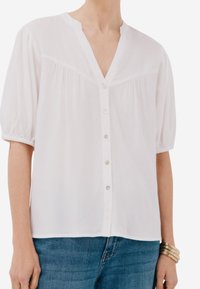 Blusa bianca a maniche corte con scollatura a V, chiusura anteriore con bottoni, dettagli arricciati e un tessuto morbido e leggero. Abbinata a jeans blu.