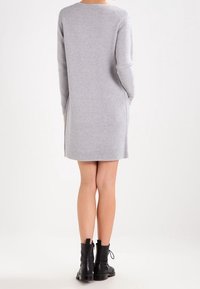 Vrouw in een lichtgrijze ribbeljurk met lange mouwen en zakken, zwarte enkelboots, staand met haar rug naar de camera op een effen witte achtergrond.