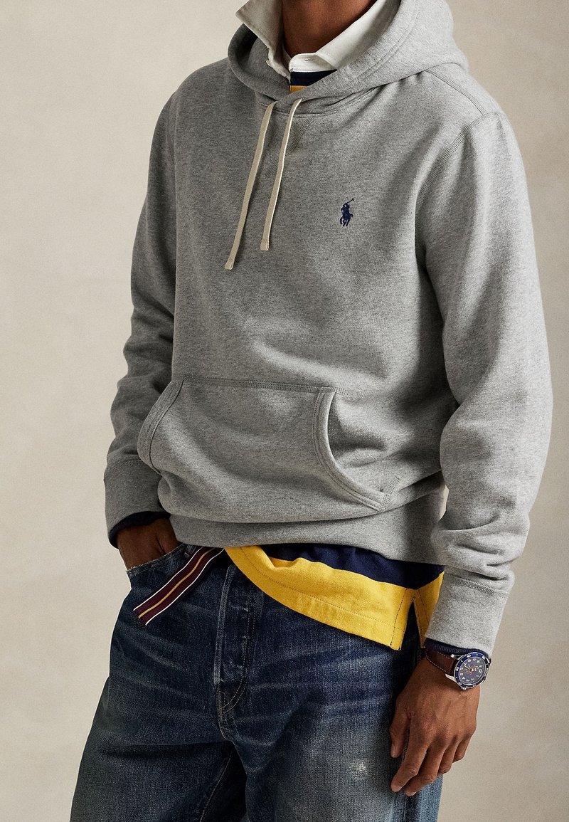 Personne portant un sweat à capuche gris sur une chemise rayée à col et un t-shirt jaune, avec un jean bleu et une montre-bracelet. Mains partiellement dans les poches.