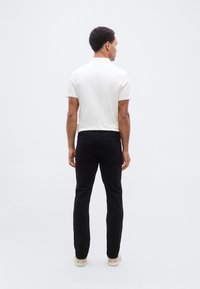 Άσπρο polo με κοντά μανίκια, φορεμένο με μαύρα τζιν και ανοιχτόχρωμα παπούτσια, από πίσω σε απλό φόντο.