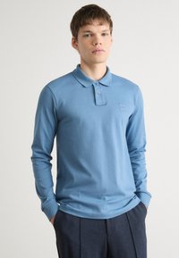 Langarm-Polohemd aus leichtem Baumwollstoff in Hellblau, mit einem Kragen, einer dreiknöpfigen Knopfleiste und einem kleinen Logo-Patch auf der Brust.