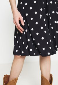 Kvinnas hand vilar nära knä-lång svart kjol med vita prickar, i kombination med beige mocka ankelboots.