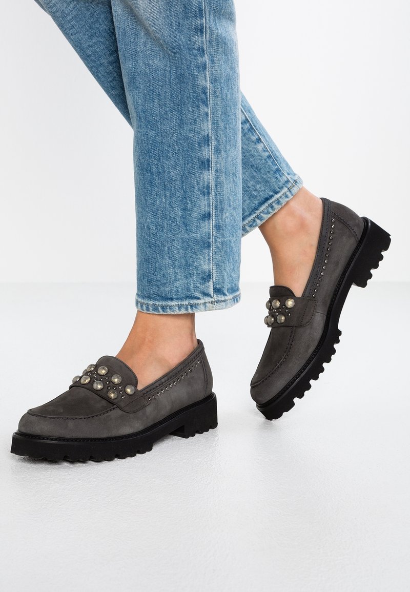 Mocassins en suède gris avec une semelle noire épaisse, présentant un cluster décoratif de clous métalliques sur le dessus et des coutures détaillées.