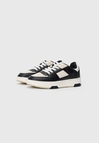 Zapatillas deportivas negras y beiges con upper texturizado, cordones blancos y suela de color crema contrastante. Presentan un diseño elegante y moderno.