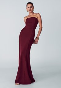 Bezramenné burgundské šaty s priliehavým korzetom a rozšírenou sukňou. Hladká, elastická tkanina s elegantným strihom. Ozdobené zlatými šperkami.