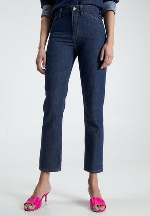 Donna che indossa jeans blu scuro a gamba dritta e sandali aperti con tacco rosa acceso su sfondo semplice.