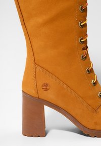 Botín de ante naranja con tacón, ojales hexagonales, cordones amarillos en contraste y un logo de árbol grabado cerca del tacón. Acabado texturizado, diseño hasta la rodilla.
