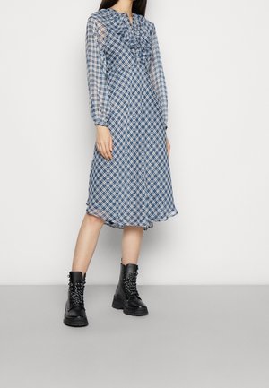 Vrouw draagt een blauw-wit geruite midi-jurk met ruches bij de halslijn en doorzichtige lange mouwen, gecombineerd met zwarte vetercombatlaarzen.