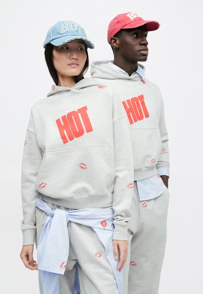 �Δύο μοντέλα φορούν ταιριαστά γκρι φούτερ με κουκούλα και παντελόνια με κόκκινο κείμενο «HOT» και στάμπες φιλιών· το ένα φορά μπλε καπέλο, το άλλο ροζ καπέλο, και οι δύο κοιτούν προς αντίθετες κατευθύνσεις.
