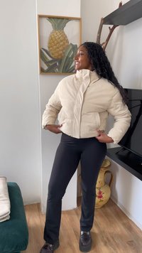 Mulher a sorrir, vestindo uma jaqueta puffer bege e calças pretas, em pé na sala de estar com uma pintura de abacaxi e um vaso decorativo.
