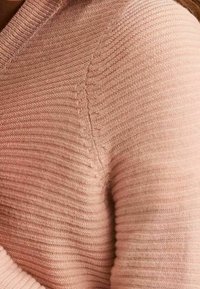 Ljust rosa stickad tröja med ett strukturerat ribbmönster och V-ringning. Funktioner inkluderar en figurnära silhuett och något upphöjda sömmar.