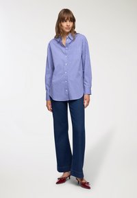 Camicia button-up a righe blu e bianche con maniche lunghe, taschino sul petto, abbinata a pantaloni a gamba larga blu scuro e scarpe con tacco rosse.