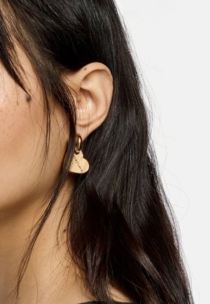 Pendiente en forma de corazón de oro con un acabado suave y texto grabado. Cuelga de un pequeño aro. El cabello cubre parcialmente la oreja.