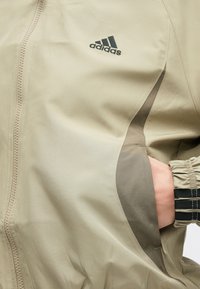 Béžová bunda Adidas so zipsom vpredu, bočnými vreckami, elastickými manžetami a kontrastnou tmavšou časťou na boku.