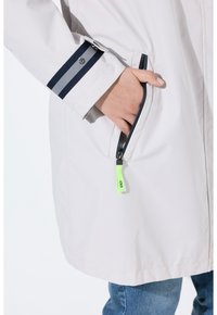 Veste légère gris clair avec une poche zippée, un poignet rayé noir et gris, et un détail de tirette zippée en néon vert.