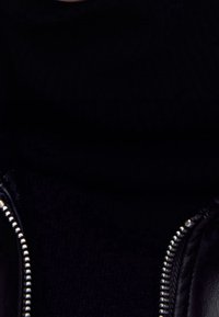 Blouson en cuir noir avec fermeture éclair argentée, superposé à un tissu en maille noire. Texture lisse et coupe ajustée.