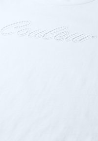 Tissu blanc avec un texte en cursive blanche pâle cousu sur la surface, créant un effet embossé subtil.