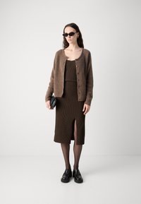 Cardigan en tricot brun sur une robe côtelée brun foncé avec une fente sur le devant. Lunettes de soleil rondes noires et chaussures à plateforme. Textures lisses, design décontracté.