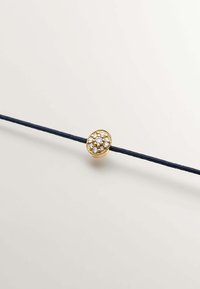 Charm oval dorado adornado con cristales claros, unido a un cordón trenzado azul marino, con bordes detallados y una textura suave.