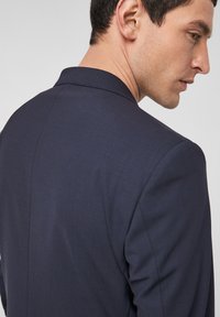 Tumedat sinise, tekstureeritud ülikonnavärk, millel on slim fit, sakiline krae ja ühe avaga taga, näidates kaasaegset ja kohandatud disaini.
