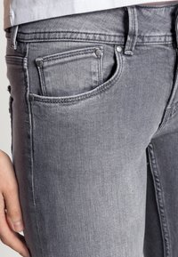 Vaqueros de mezclilla gris con un ajuste slim, bolsillos clásicos y acentos de hardware metálico. La tela tiene una textura suave con costuras sutiles.