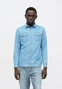 Gaiši zilas džinsa krekls ar garām piedurknēm, pogājamu priekšpusi, divām krūšu kabatām un izliekta apakša, valkāts kopā ar gaiši zilām džinsu biksēm.