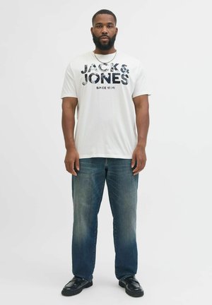 Valge lühikeste varrukatega t-särk, millel on "JACK & JONES" grafika, kombineeritud siniste teksadega. Inimene seisab püsti ja kannab musti kingi.