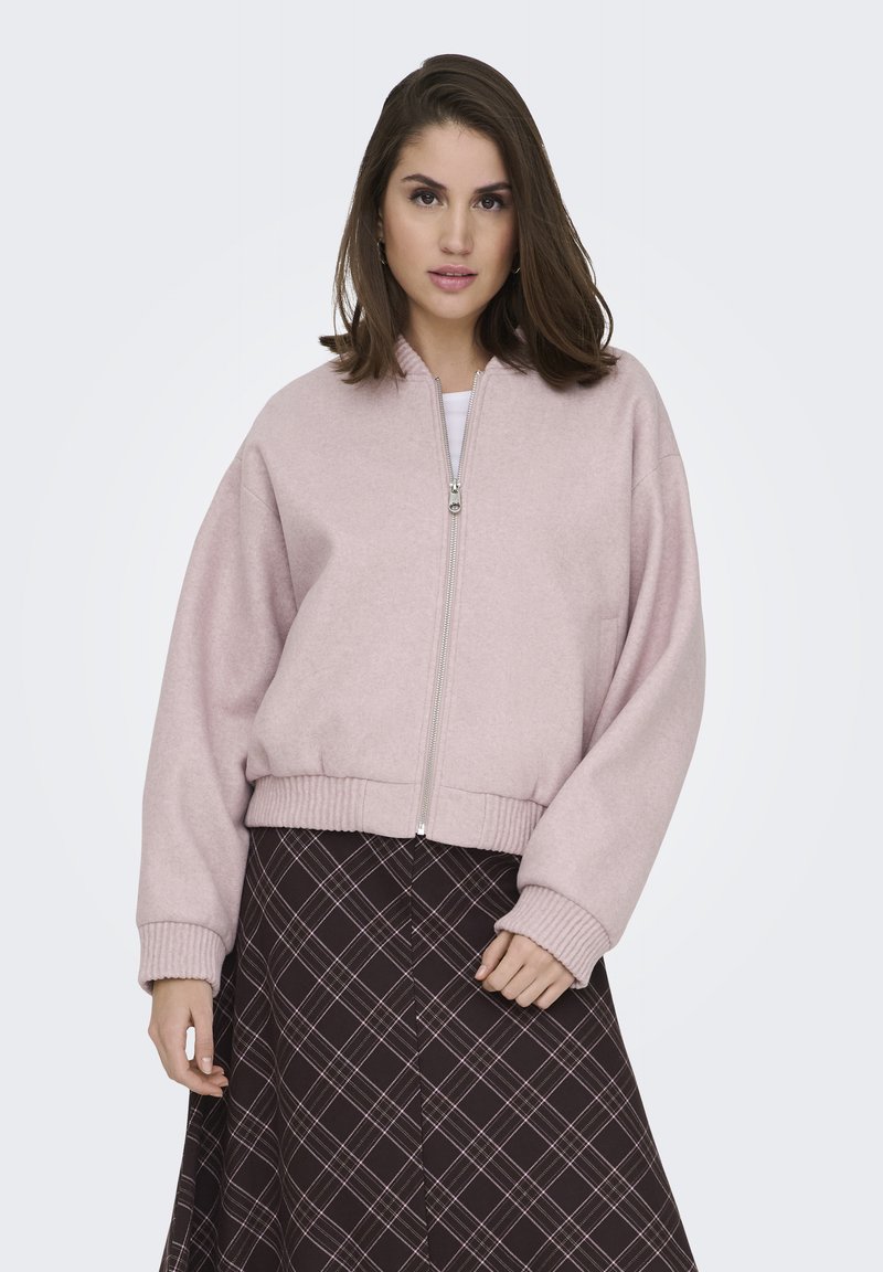 Veste bomber en polaire rose avec une fermeture éclair à l'avant, des poignets et un ourlet côtelés. Portée sur une jupe marron à motifs avec des lignes diagonales.