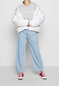 Persona vistiendo una chaqueta acolchada blanca sobre una camiseta gris de Calvin Klein, jeans de pierna ancha azul claro y zapatillas rojas de caña alta.