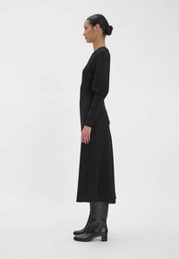 Robe longue noire à coupe ajustée avec manches bouffantes, texture lisse et longueur midi, associée à des bottes noires montantes jusqu'aux genoux.
