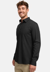 Camisa negra de manga larga con botones, cuello clásico, corte recto y textura suave, llevada con pantalones de color claro.