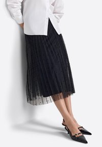 Schwarzer plissierter Tüllrock mit Polka-Dot-Muster; getragen mit einem weißen Hemd mit Knöpfen und schwarzen Slingback-Schuhen mit spitz zulaufender Spitze.