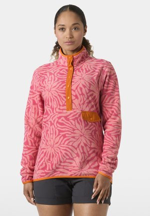 Kobieta ubrana w różowy polarowy sweter z wzorami, z pomarańczowymi lamówkami i guzikami, w czarnych szortach, stojąca na jednolitym tle.