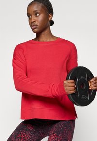 Felpa rossa a maniche lunghe con collo rotondo, abbinata a un peso in ghisa nero e leggings a fantasia con tonalità di rosso e nero.