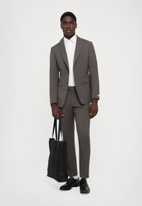 Costum gri, croit cu rever, asortat cu o cămașă albă și pantofi negri. Modelul ține o geantă tote neagră. Design simplu, modern.