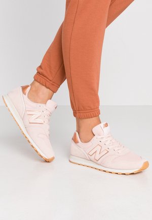 Personne portant des baskets New Balance rose clair avec des semelles beige et un pantalon de jogging couleur rouille, debout sur un sol blanc.