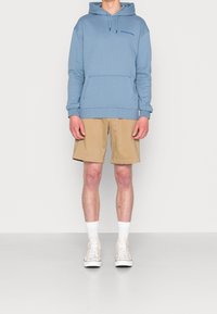 Sudadera azul claro con bolsillo frontal y logo impreso, combinada con pantalones cortos beige. El modelo lleva calcetines blancos y zapatillas claras.