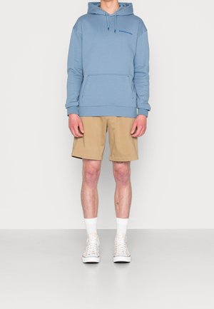 Sudadera azul claro con bolsillo frontal y logo impreso, combinada con pantalones cortos beige. El modelo lleva calcetines blancos y zapatillas claras.