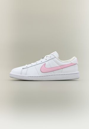 Witte lage sneaker met roze Nike-swoosh en veters, gestructureerde witte rubberen zool, tentoongesteld op een neutrale lichte achtergrond.