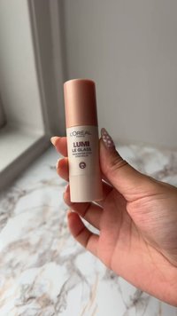 L'Oréal Lumi Le Glass highlighterstick in een cilindrische beige verpakking met een roségouden dop, met daarop tekst en een logo in donkerpaars.
