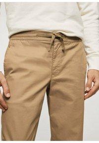 Pantalon beige à cordon de serrage avec une ceinture élastique, doté de poches latérales et d'une texture tissée lisse.