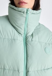Giubbotto puffer verde menta chiaro con colletto alto, zip frontale e dettaglio del logo arrotondato. Texture morbida e design trapuntato visibile.
