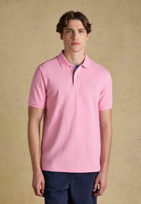 Jeune homme portant un polo à manches courtes rose clair et un pantalon bleu marine, debout devant un fond beige uni.