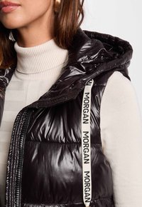 Gilet in piuma nero lucido con cappuccio, dotato di un'etichetta bianca del marchio sul lato e di un dolcevita crema a coste sotto.