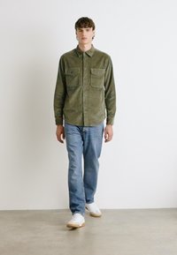 Olijfgroene ribfluwelen overhemden met twee borstzakken, met knopen, gecombineerd met lichtblauwe jeans en witte sneakers, staand tegen een neutrale achtergrond.