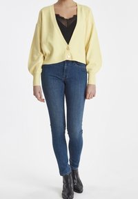 Gul kortärmad cardigan med V-ringning, knäppning och puffärmar, i kombination med åtsittande mörkblå jeans och svarta ankelboots.