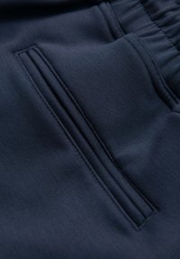 Gros plan d'une poche de vêtement bleu marine avec une fente verticale et des coutures visibles, fixée à une section de ceinture élastique.